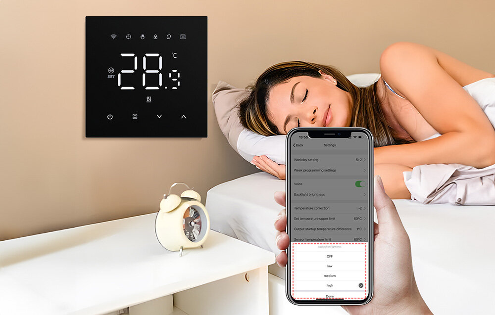Na ścianie wisi czarny panel z wyświetlaczem pokazującym temperaturę 28°C oraz ikonami. Osoba trzyma w ręku smartfon z aplikacją do sterowania termostatem, a w tle widać śpiącą kobietę i budzik na stoliku.