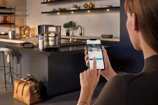 SIEMENS serii EQ900 Plus - kobieta trzymająca smartfon, w tle ekspres na wyspie kuchennej