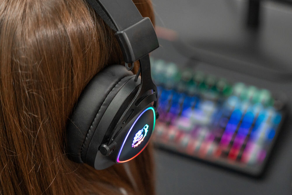 Słuchawki MAD DOG GH200K Nauszne Zbliżenie na osobę w czarnych słuchawkach gamingowych Mad Dog GH200 z podświetleniem RGB na nauszniku, siedzącą przed kolorową klawiaturą mechaniczną.