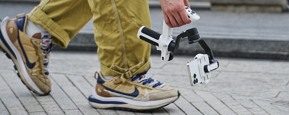 Mężczyzna trzyma gimbal FeiyuTech Scorp mini P. Nagrywa telefonem materiał wideo, prowadząc sprzęt tuż nad ziemią.