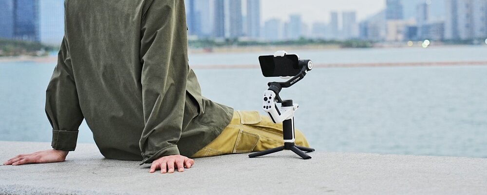 Mężczyzna siedzi na betonie nad brzegiem wody. Obok stoi biały gimbal FeiyuTech z zamontowanym smartfonem.
