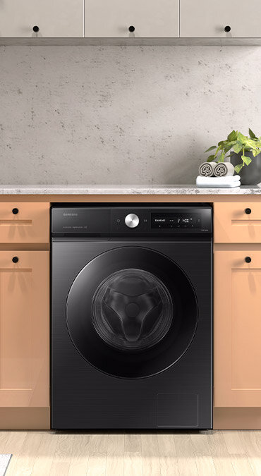 Czarna pralka w zabudowie kuchennej z widocznym panelem sterowania i okrągłym pokrętłem. Na blacie obok pralki stoi roślina w doniczce.