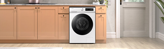 Pralka w kolorze białym z czarnym panelem i okrągłym drzwiczkami, wkomponowana w zabudowę kuchenną z jasnobrązowymi szafkami. Na blacie znajduje się zlew i kilka przedmiotów kuchennych.