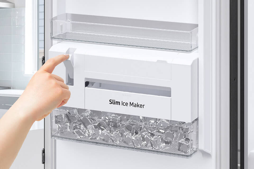 Wbudowana w drzwi kostkarka nie zabiera cennego miejsca wewnątrz Osoba dotyka panelu urządzenia do robienia lodu z napisem \'Slim Ice Maker\'. W dolnej części widoczne są kostki lodu.