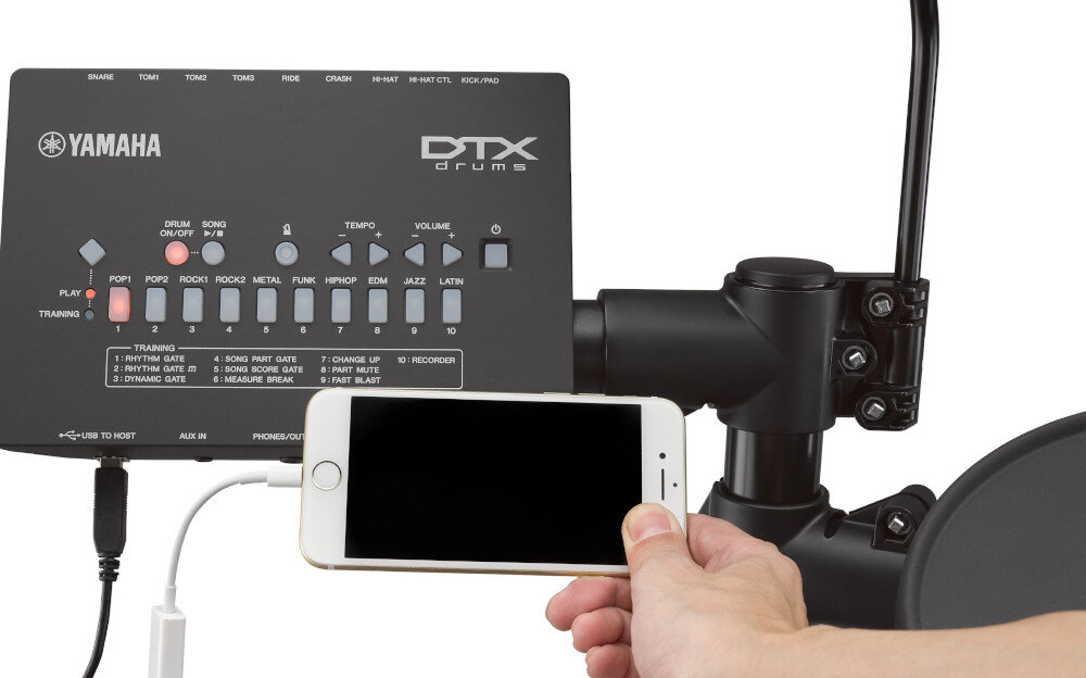 Panel sterowania perkusji elektronicznej z logo YAMAHA i DTX drums, z różnymi przyciskami i wskaźnikami. Osoba trzyma smartfon podłączony do panelu za pomocą kabla.