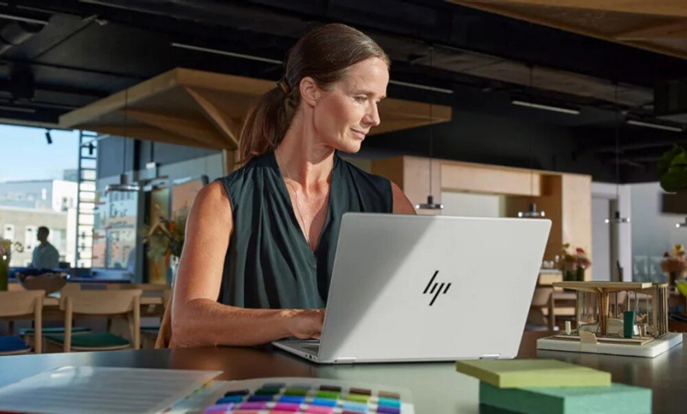 Laptop HP EliteBook 1040 G10 Kobieta siedząca przy stole w przestronnym biurze, pracująca na srebrnym laptopie. W tle widać drewniane elementy wystroju, lampy wiszące i inne osoby w biurze, opis