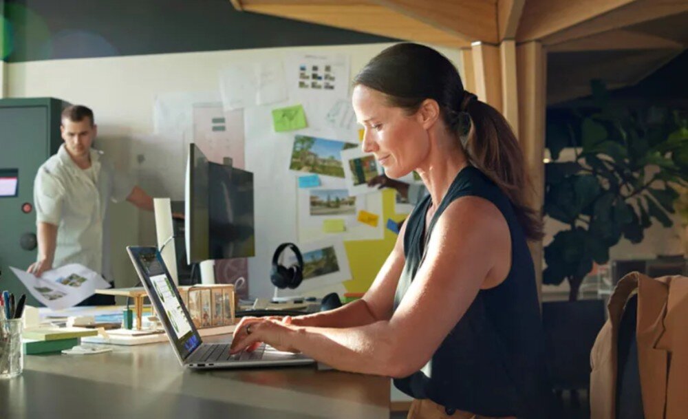 Laptop HP EliteBook 1040 G10 Kobieta siedząca przy biurku w biurze, pracująca na laptopie. W tle mężczyzna przy tablicy z projektami architektonicznymi, podświetlenie klawiatury, touchpad, czytnik linii papilarnych