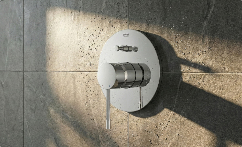 BATERIA WANNOWA PODTYNKOWA GROHE ESSENCE Zdjęcie przedstawia chromowaną baterię prysznicową zamontowaną na ścianie z ciemnych, matowych płytek. Światło podkreśla połysk metalu, tworząc elegancki efekt