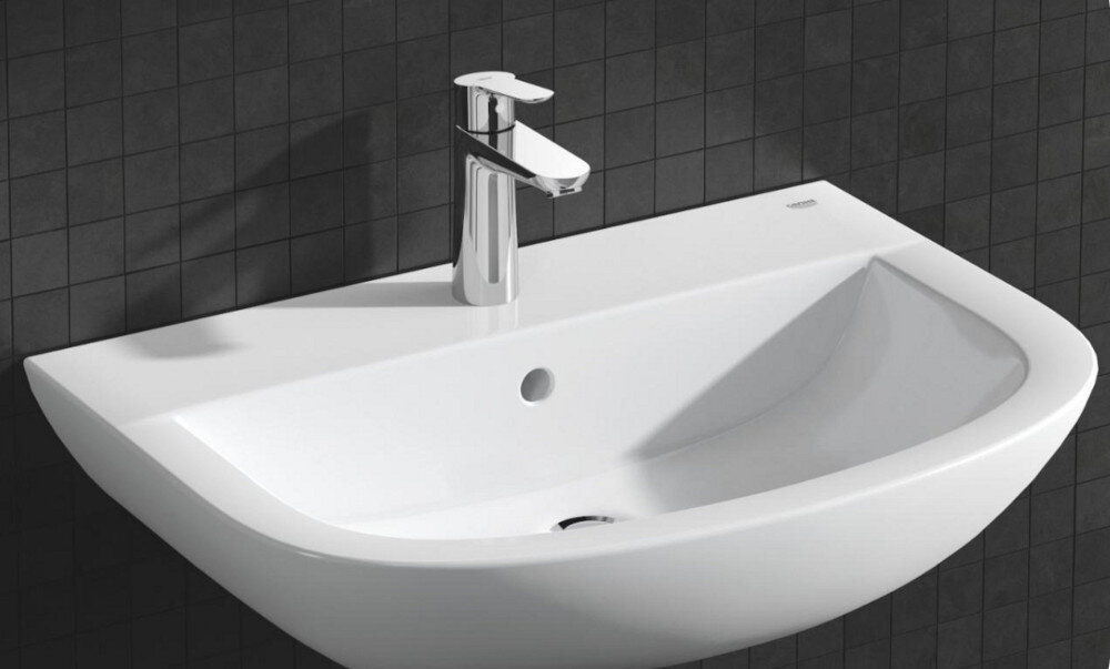 BATERIA UMYWALKOWA GROHE BAUEDGE ROZMIAR M 23758000 CHROM Zdjęcie przedstawia białą umywalkę o asymetrycznej formie, zamontowaną na tle ciemnych płytek ściennych. Na umywalce znajduje się chromowana bateria jednouchwytowa, podkreślająca minimalistyczny charakter aranżacji