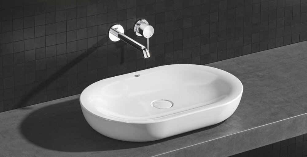 BATERIA UMYWALKOWA 2-OTWOROWA GROHE ESSENCE Zdjęcie przedstawia elegancką baterię ścienną o chromowanym wykończeniu, z której wypływa strumień wody. Minimalistyczny design podkreśla jasne tło o delikatnej strukturze