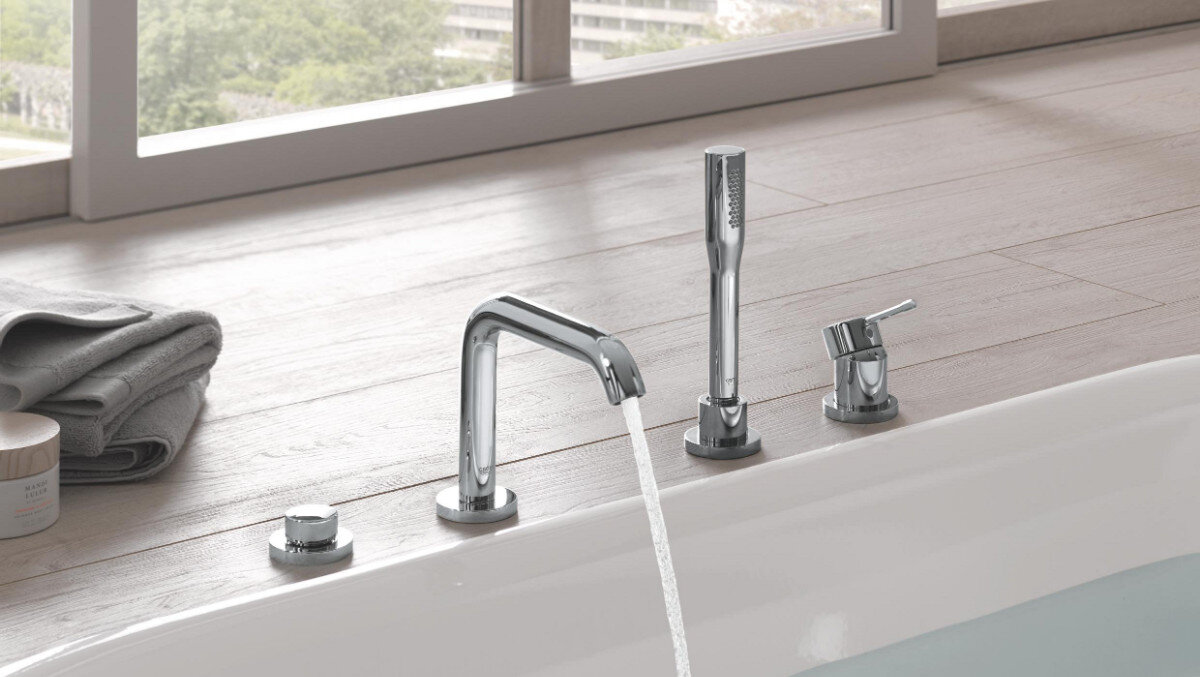 Na zdjęciu widać chromowaną baterię wannową z wylewką, słuchawką prysznicową i pokrętłem. W tle znajduje się drewniana powierzchnia oraz okno z widokiem na zieleń.