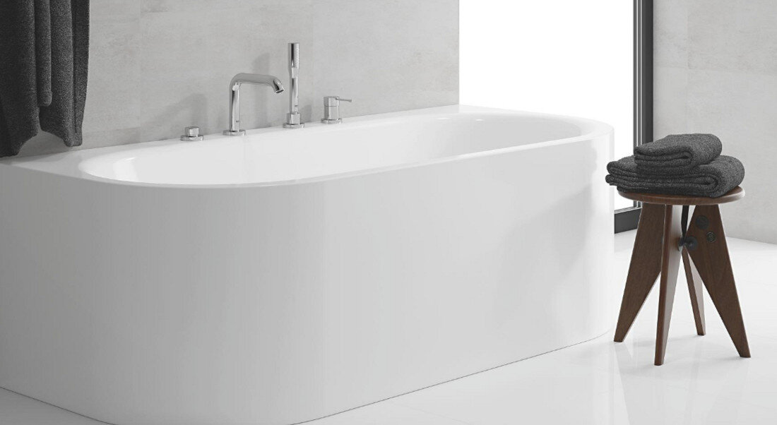 Na zdjęciu znajduje się nowoczesna, biała wanna wolnostojąca z chromowaną baterią wannową. Obok wanny stoi drewniany stołek z ciemnoszarymi ręcznikami.