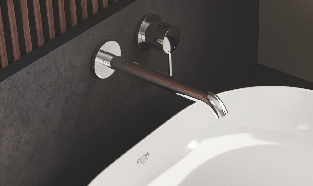 BATERIA UMYWALKOWA 2-OTWOROWA GROHE ESSENCE Zdjęcie przedstawia elegancką baterię ścienną o chromowanym wykończeniu, z której wypływa strumień wody. Minimalistyczny design podkreśla jasne tło o delikatnej strukturze
