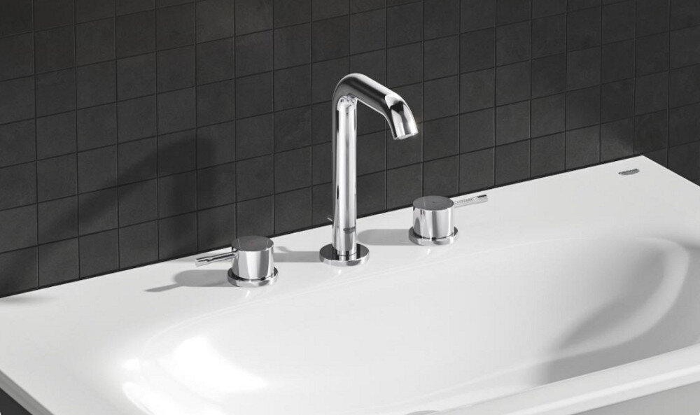 Chromowana bateria umywalkowa z dwoma pokrętłami na białej umywalce, na tle czarnej, kafelkowej ściany.