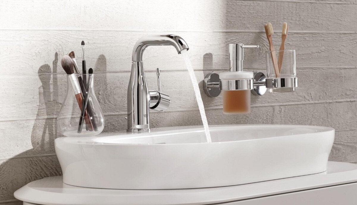 BATERIA UMYWALKOWA GROHE ESSENCE ROZMIAR M 23462001 CHROM Na zdjęciu widać chromowaną baterię umywalkową z bieżącą wodą, otoczoną akcesoriami łazienkowymi, takimi jak pędzle do makijażu i dozownik mydła