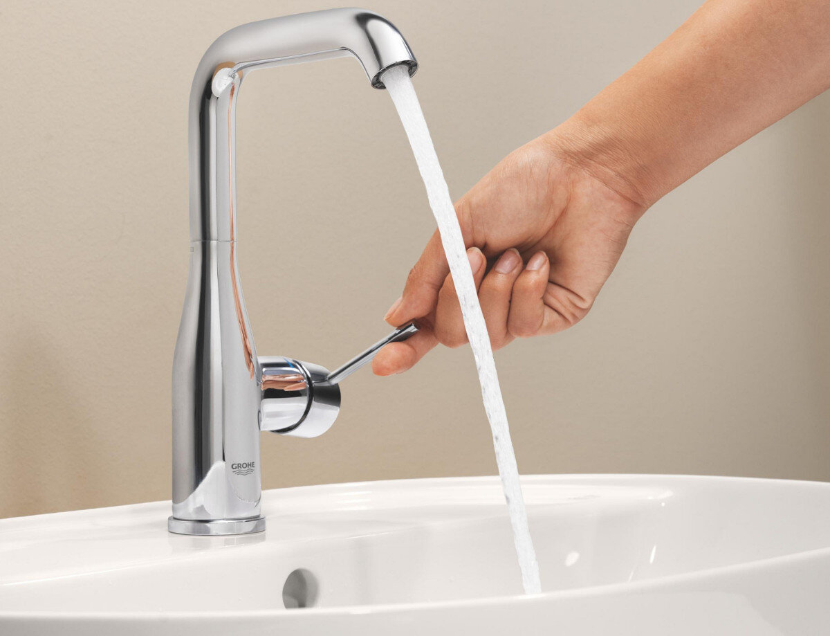 BATERIA UMYWALKOWA GROHE ESSENCE ROZMIAR L 23541001 CHROM Na zdjęciu widać chromowaną baterię umywalkową, z której wypływa strumień wody, a obok pokazano dłoń regulującą przepływ za pomocą bocznej dźwigni. Ujęcie podkreśla wygodę obsługi oraz precyzję sterowania wodą w nowoczesnej armaturze estetyka GROHE Essence chromowana powłoka oszczędność wody płynna obsługa