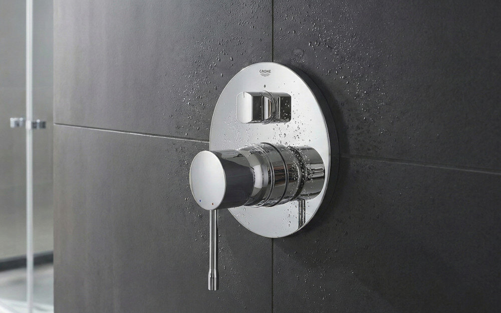 BATERIA WANNOWA PODTYNKOWA GROHE ESSENCE Zdjęcie przedstawia chromowaną baterię prysznicową zamontowaną na ścianie z ciemnych, matowych płytek. Światło podkreśla połysk metalu, tworząc elegancki efekt
