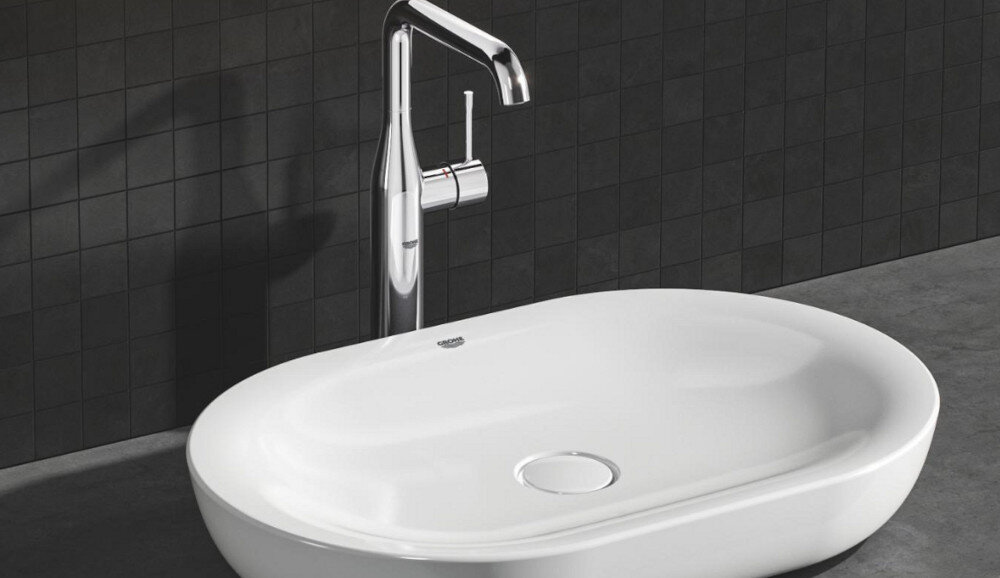 BATERIA UMYWALKOWA GROHE ESSENCE ROZMIAR XL 32901001 CHROM Zdjęcie przedstawia chromowaną baterię umywalkową zamontowaną przy owalnej, białej umywalce nablatowej na tle ciemnych płytek ściennych. Minimalistyczna forma armatury podkreśla elegancki charakter aranżacji łazienki