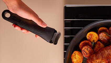 Osoba trzyma czarny uchwyt patelni z widocznym logo \'Tefal\', obok znajduje się patelnia z pieczonymi ziemniakami i mięsem.