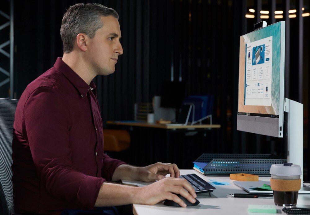 Komputer HP EliteOne 840 G9 Mężczyzna w bordowej koszuli siedzi przy biurku i pracuje na komputerze typu All-in-One, w tle ciemne wnętrze biura, opis