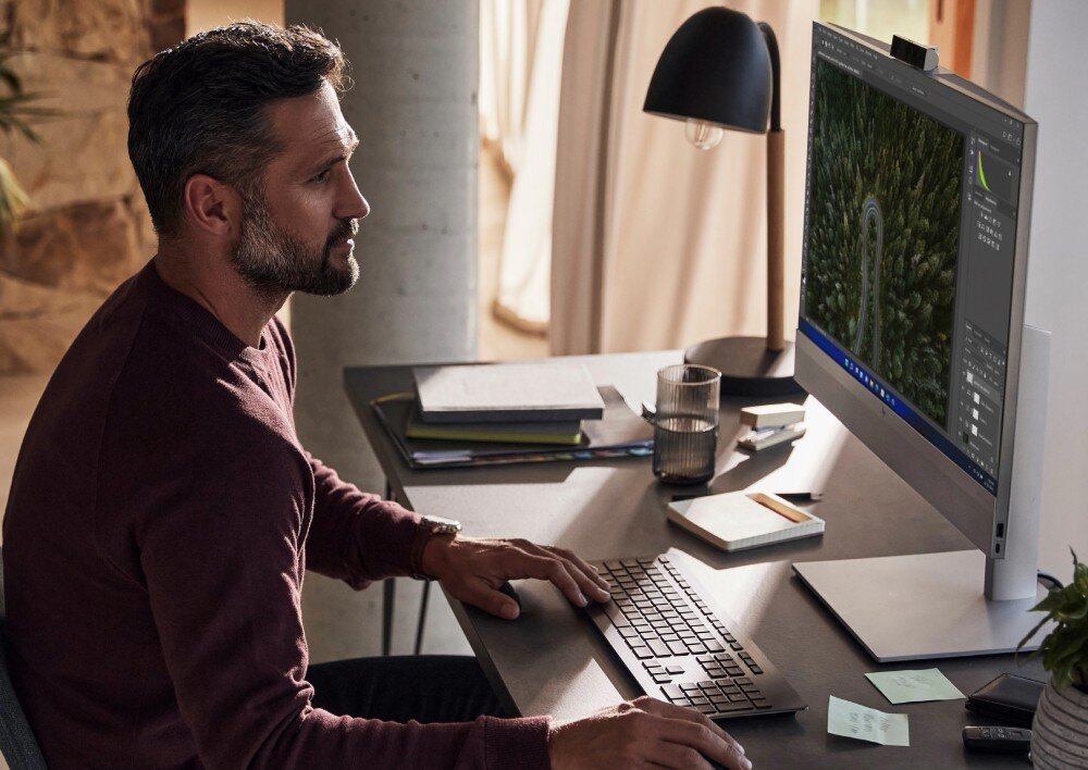 Komputer HP EliteOne 870 G9 Mężczyzna pracuje przy biurku, patrząc na ekran komputera typu all-in-one z wyświetlonym zdjęciem lasu, procesor, rdzenie, pamięć, dysk, wydajność