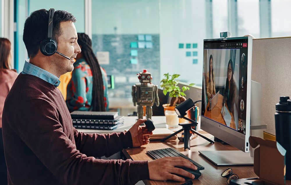 Komputer HP EliteOne 870 G9 Mężczyzna w słuchawkach z mikrofonem rozmawia online przy biurku z komputerem typu all-in-one, w tle kilka osób i dekoracyjny robot, zabezpieczenia, ochrona plików, funkcje