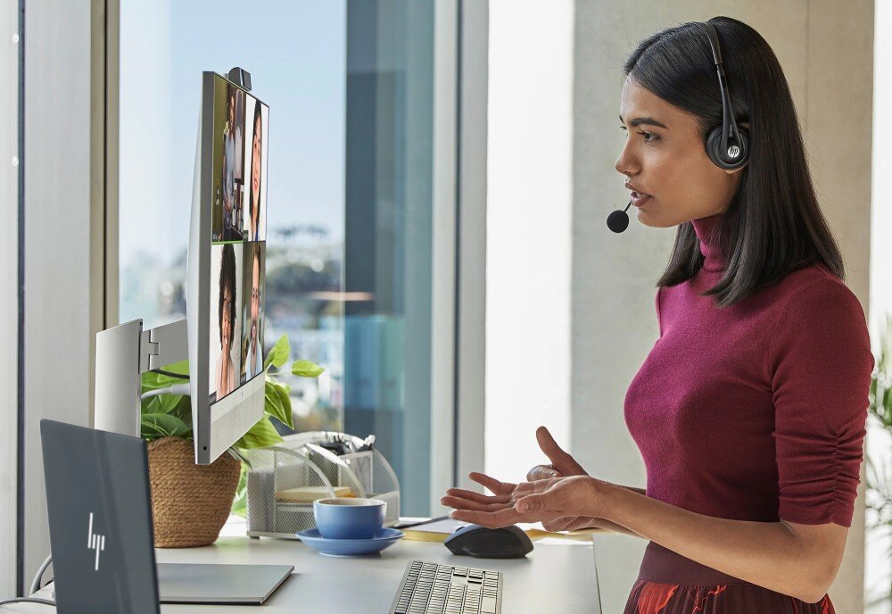 Komputer HP EliteOne 870 G9 Kobieta w słuchawkach z mikrofonem prowadzi wideokonferencję przy biurku z komputerem typu all-in-one, kamera, głośniki, dźwięk, komunikacja