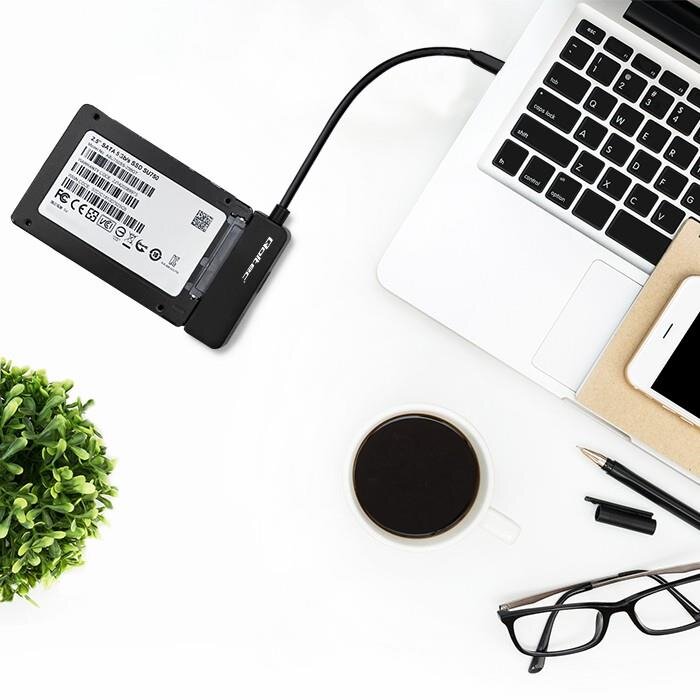 Na białym biurku znajduje się laptop z podłączonym kablem adaptera do dysku SSD, obok leży filiżanka kawy, okulary, długopis i telefon na notesie. W tle widoczna jest zielona roślina.