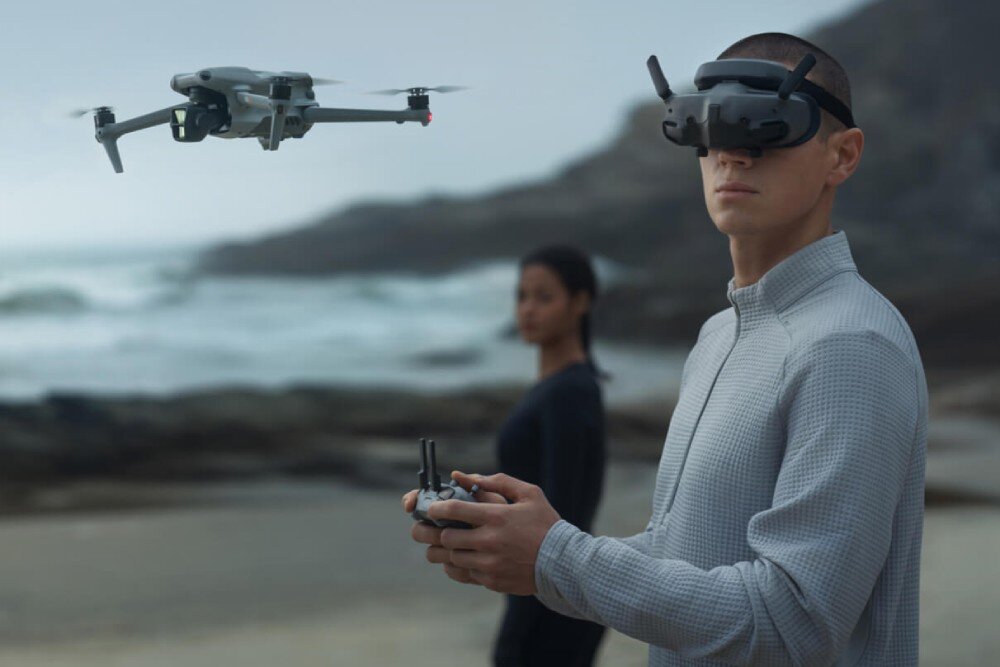 Gogle DJI Goggles 3 Zbliżenie na osobę w goglach trzymającą kontroler, w tle unosi się dron nad brzegiem morza, obok stoi kobieta