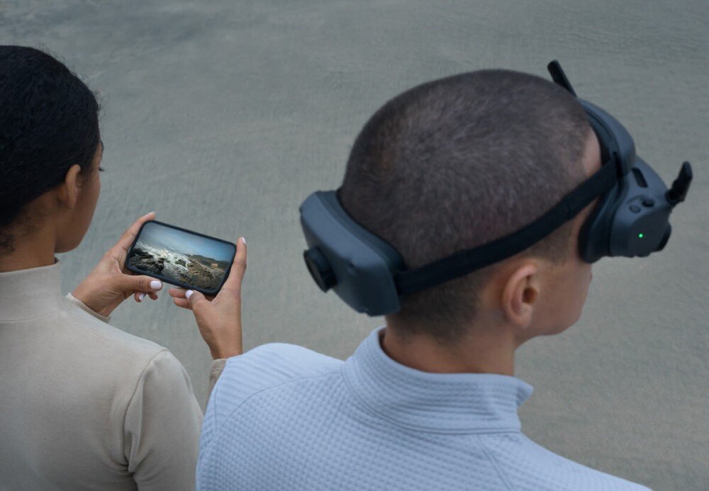 Gogle DJI Goggles 3 Widok z góry na osobę w goglach, obok kobieta trzyma smartfon z widokiem skalistego wybrzeża