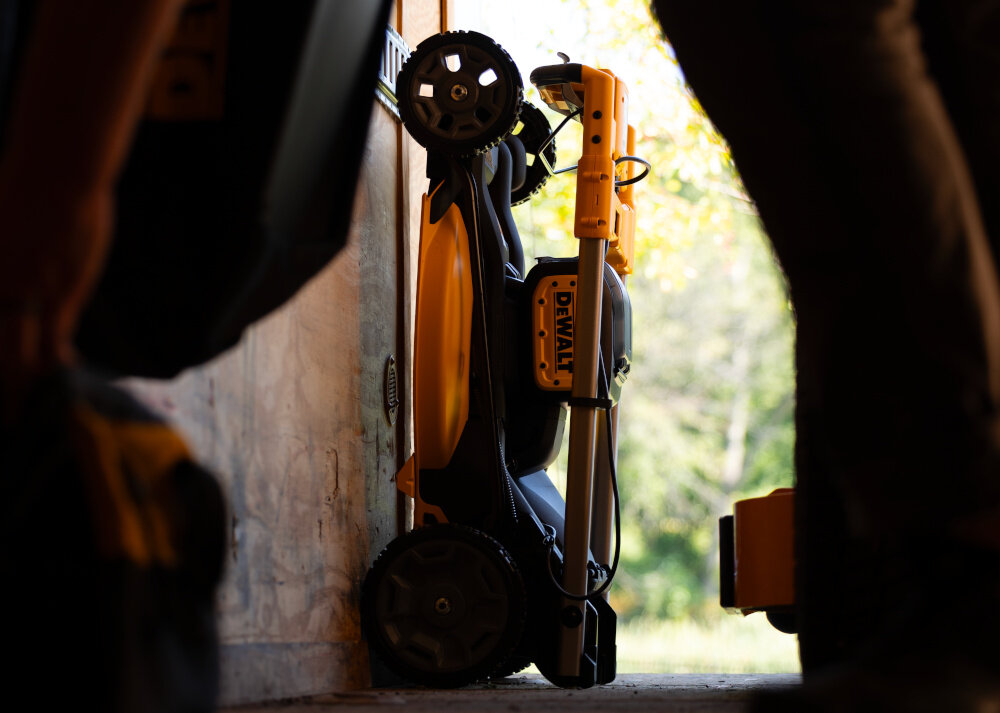 Kosiarka akumulatorowa DEWALT DCMWSP156N-XJ z napędem szopa garaż złożona kosiarka stoi oparta o ścianę wokół inne narzędzia do regularnego koszenia trawników praca bez przewodu ułatwia manewrowanie skraca czas koszenia zmniejsza wysiłek fizyczny