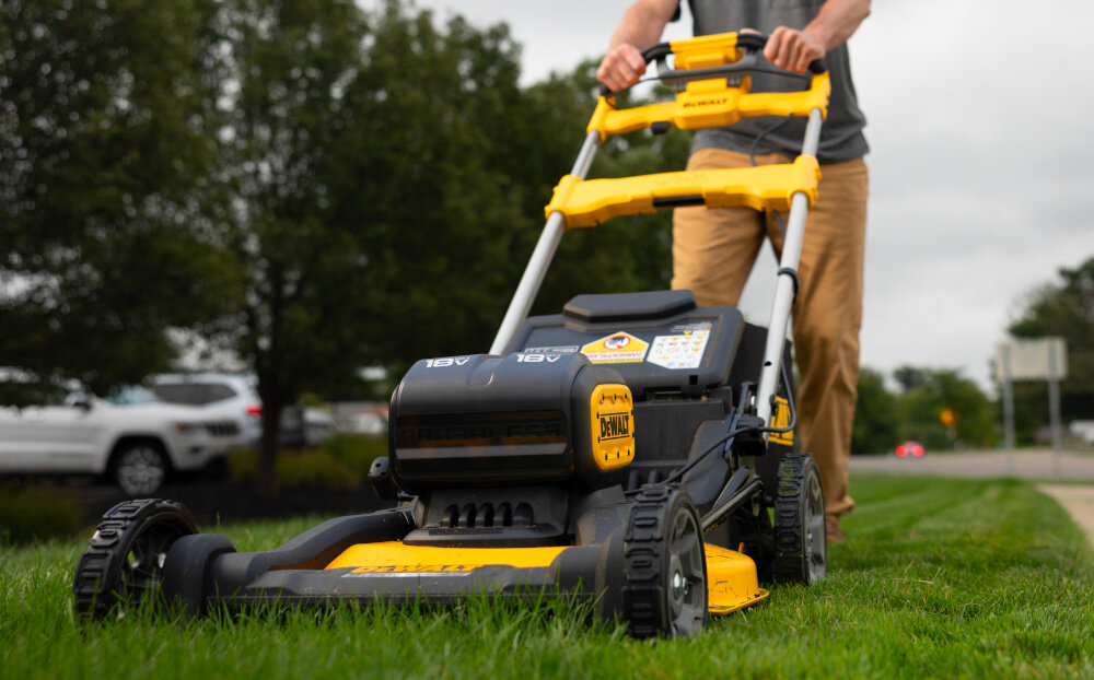 Kosiarka akumulatorowa DEWALT DCMWSP156N-XJ z napędem ogród trawnik osoba kosi trawę w tle niebo samochody drzewa silnik indukcyjny bezszczotkowy wysoka wydajność przy niższym zużyciu energii i dłuższy czas pracy na jednym ładowaniu do trawników o powierzchni około do 1340 m² zabezpieczenie przed przypadkowym uruchomieniem