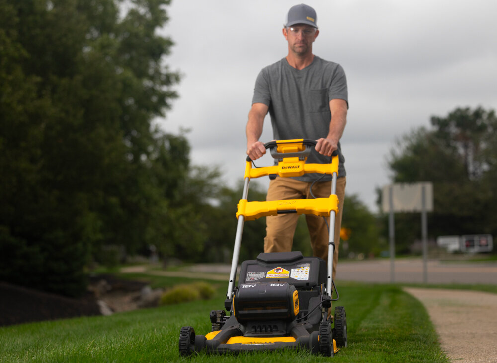 Kosiarka akumulatorowa DEWALT DCMWSP156N-XJ z napędem ogród trawnik osoba kosi trawę w tle drzewa ulica niebo napęd na tylne koła regulowana prędkość jazdy zmniejsza zmęczenie poprawia komfort pracy