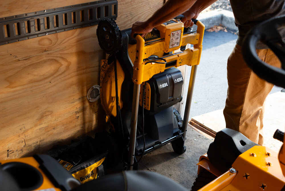 Kosiarka akumulatorowa DEWALT DCMWSP156N-XJ z napędem szopa garaż złożona kosiarka stoi na podłodze osoba trzyma dłonie na kosiarce waga około 32 kg składany uchwyt obsługiwany jednym przyciskiem lepsza organizacja przestrzeni