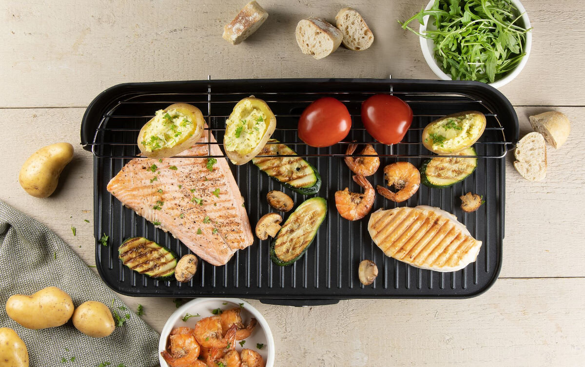 Grill elektryczny DOMO DO9260G, szybkie, równocześnie, zdejmowana, blacha, Widok z góry na grill DOMO DO9260G podczas pracy – grillujące się ryby, warzywa, krewetki i ziemniaki, dodatki rozłożone wokół urządzenia.