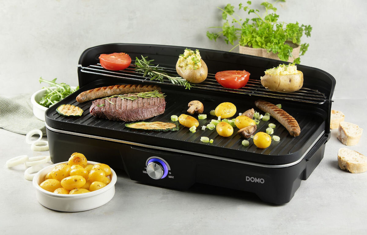 Grill elektryczny DOMO DO9260G, pielęgnacja, jakośc, powłoka, resztki, Grill elektryczny DOMO DO9260G wypełniony różnorodnymi produktami – widok od przodu na grillujące się warzywa, kiełbasy i stek, w tle zioła oraz dodatki.