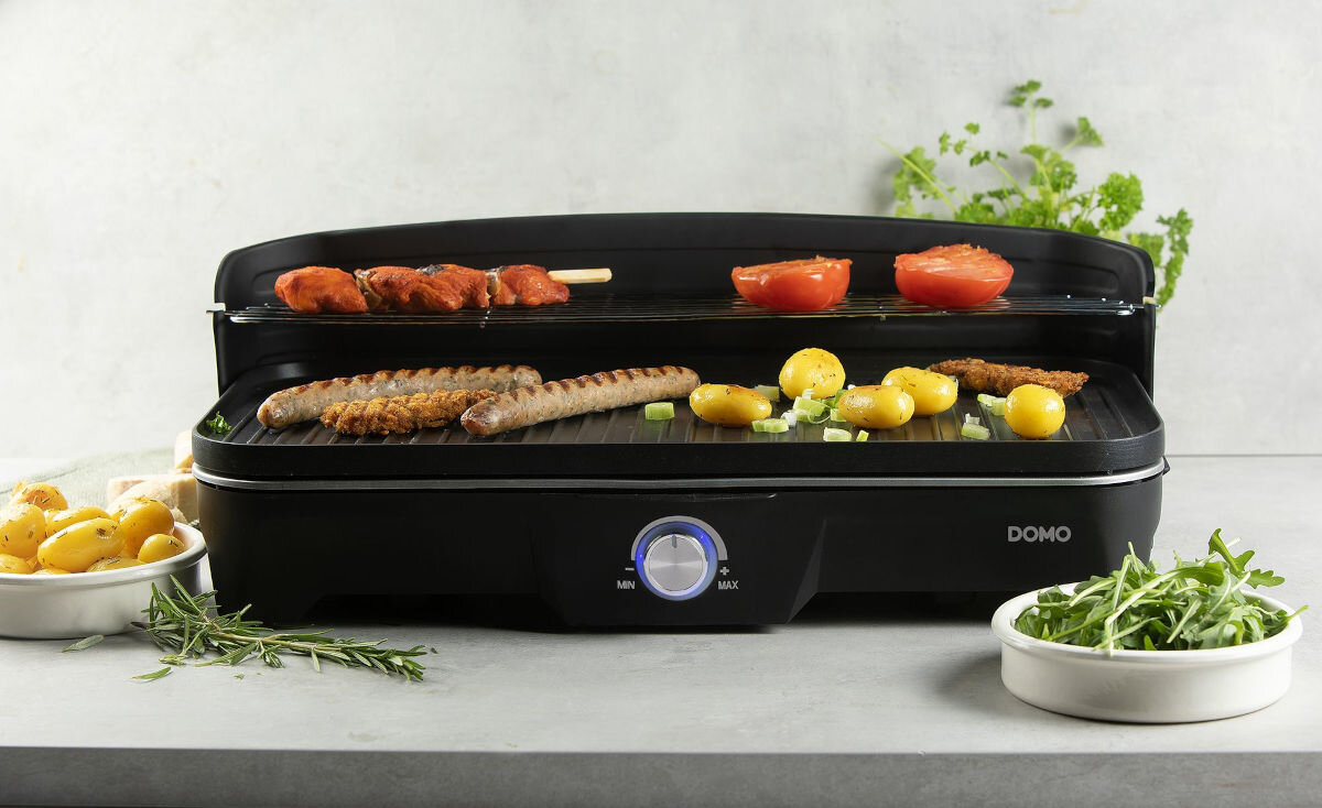 Grill elektryczny DOMO DO9260G, przekąski, sezonowe, elektryczne, spotkania, letnie, Grill DOMO DO9260G w trakcie przygotowywania różnych potraw, widok z przodu na kiełbaski, pomidory i ziemniaki, w tle świeże zioła i warzywa.