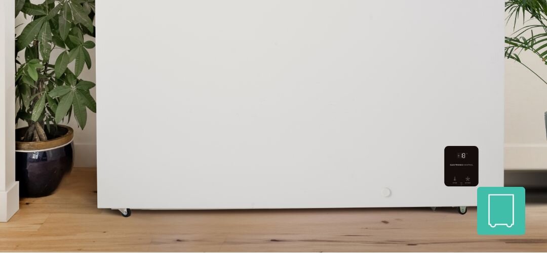 ZAMRAŻARKA GORENJE FHC42EAW Dolna część białej zamrażarki skrzyniowej stojącej na drewnianej podłodze, z widocznymi kółkami jezdnymi, panelem sterowania w prawym dolnym rogu oraz doniczkową rośliną po lewej stronie kółka transportowe łatwe przesuwanie mobilna zamrażarka wygodne przestawianie