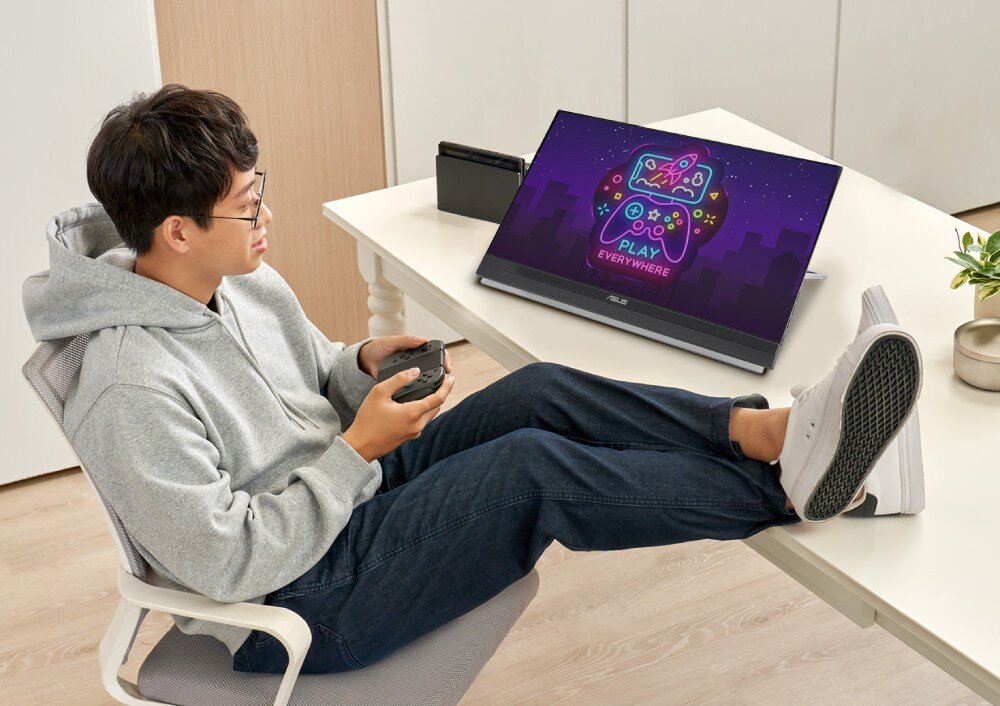 Monitor ASUS ZenScreen MB229CF Mężczyzna siedzi przy biurku, grając na konsoli podłączonej do monitora ustawionego na stole. Na ekranie widać kolorową grafikę z napisem 'Play Everywhere', głośniki, dźwięk, audio