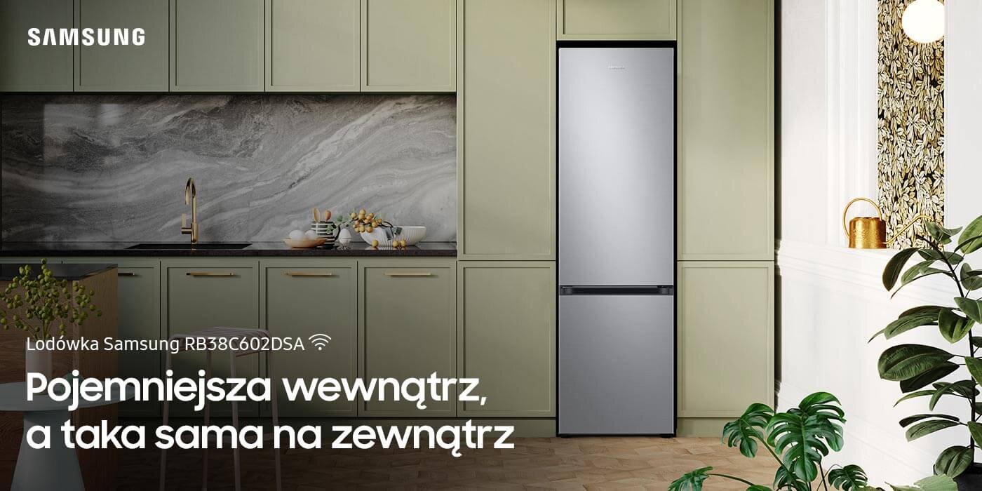 Lodówka Samsung Grand+ RB38C602DSA w kolorze metalicznego grafitu wstawiona w elegancką zabudowę kuchenną Kuchnia z lodówką Samsung RB38C602DSA w metalicznym grafitowym kolorze, otoczona zielonymi szafkami i dekoracjami. Tekst na obrazie: \'Lodówka Samsung RB38C602DSA Pojemniejsza wewnątrz, a taka sama na zewnątrz\'.