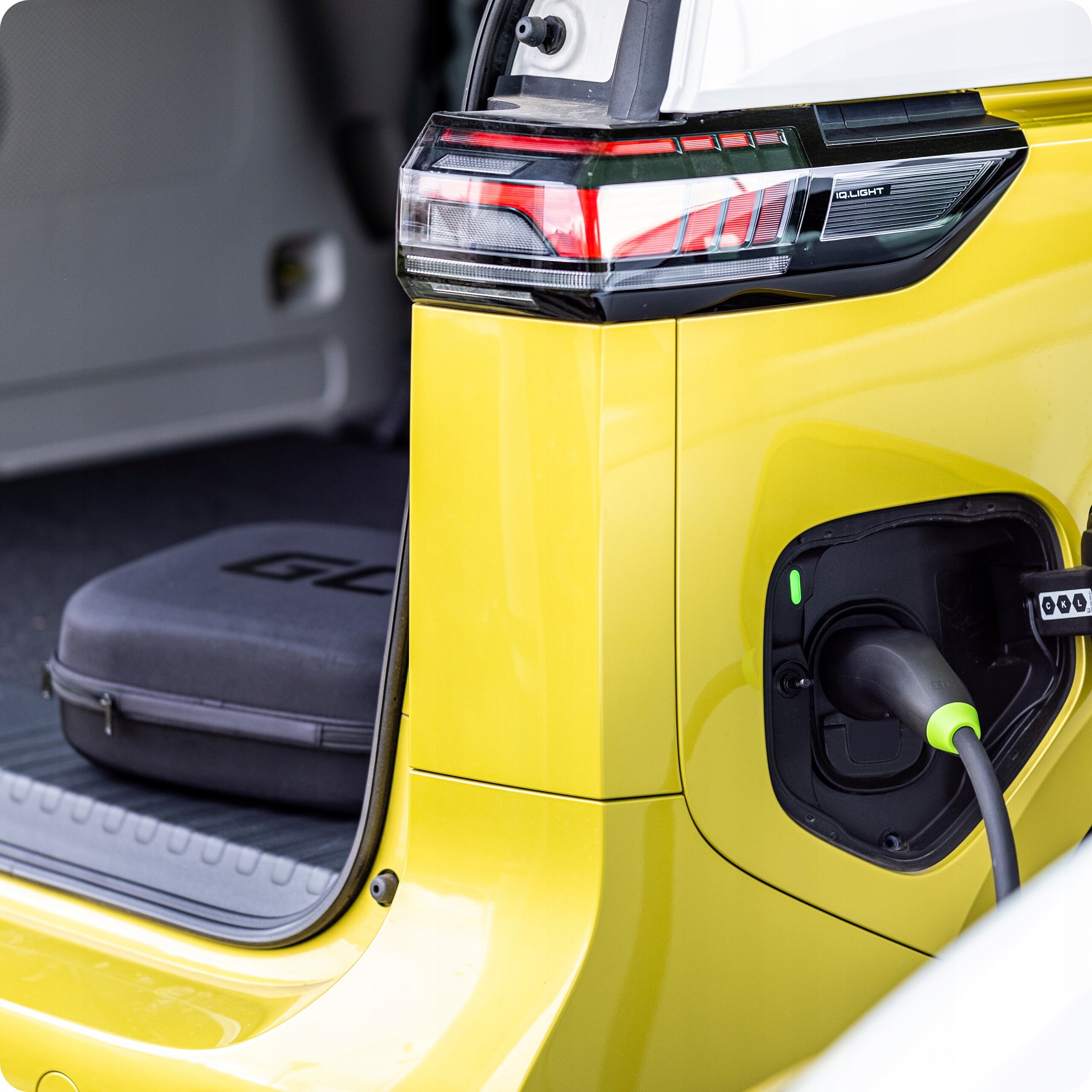 Żółty samochód elektryczny z podłączonym kablem do ładowania. W tle widoczna jest otwarta przestrzeń bagażnika z czarną torbą z napisem \'GL\'.