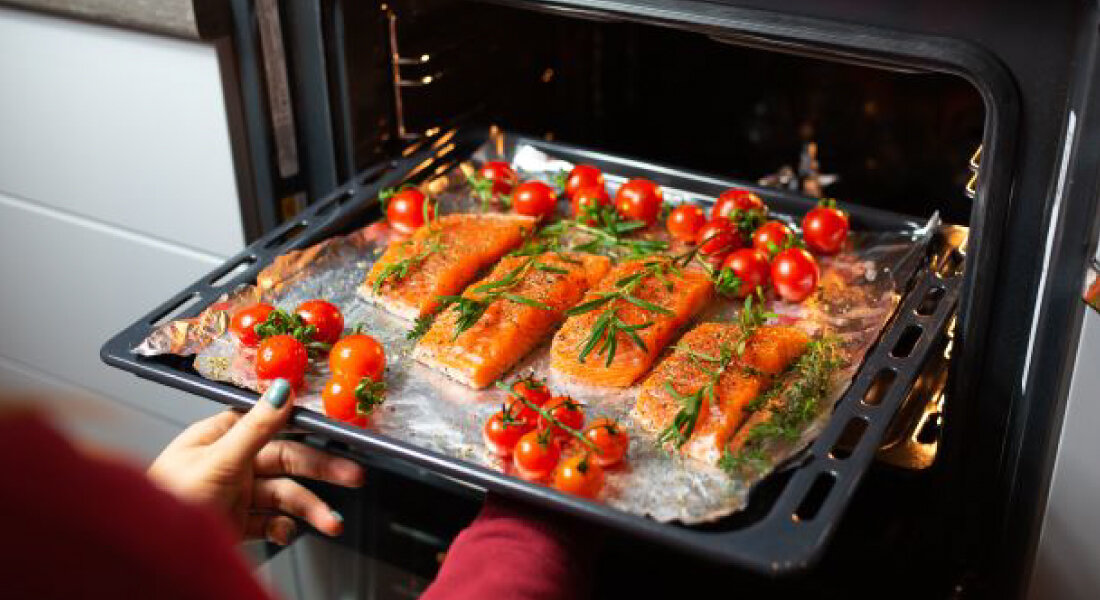 Piekarnik ETA Basic 178590000 Osoba wysuwa blachę z piekarnika z kawałkami ryby i pomidorkami koktajlowymi. Potrawa ułożona jest na papierze do pieczenia 8 programów ruszt grillowy dwie blachy klasyczne drabinki