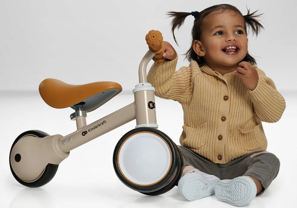 Kinderkraft rowerek biegowy CUTIE LIGHT BEIGE pierwsza jazda beżowy rowerek siodełko gumowe uchwyty czarno białe koła obok siedzi maluch i trzyma kierownicę