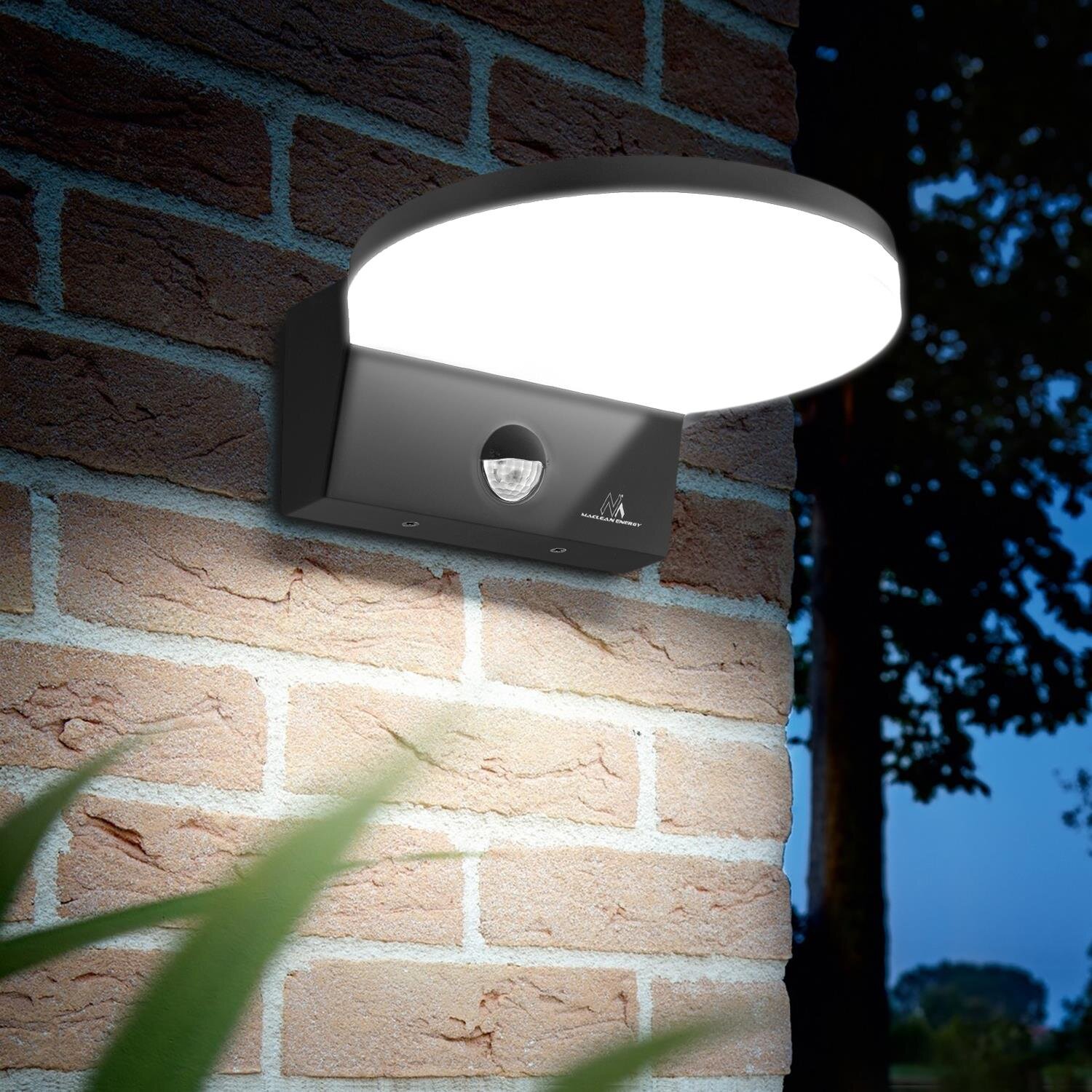 Na obrazie widoczna jest lampa LED zamontowana na ceglanej ścianie, emitująca jasne światło. Na obudowie lampy znajduje się czujnik ruchu oraz logo producenta.