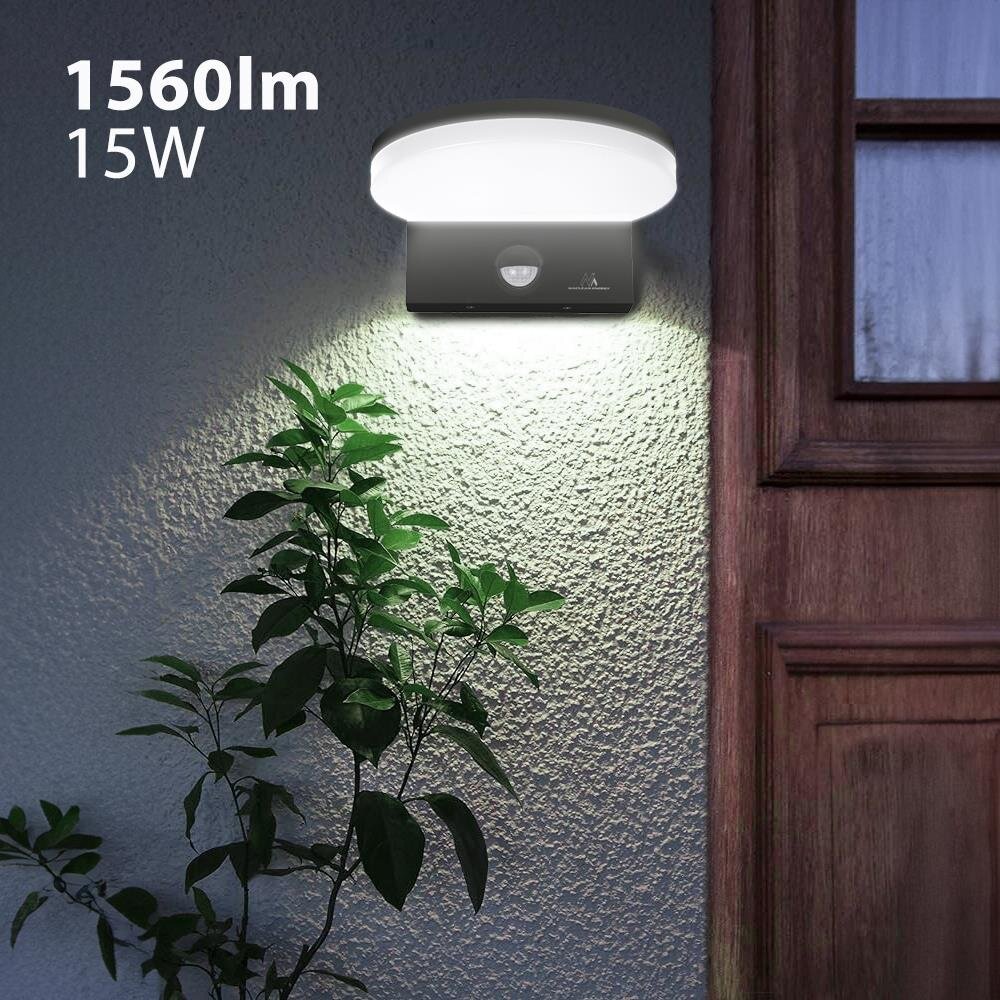 Na ścianie zamontowana jest lampa LED z czujnikiem ruchu, emitująca białe światło. Obok widoczny jest fragment drewnianych drzwi oraz roślina, a na obrazie znajduje się tekst: \'1560lm 15W\'.