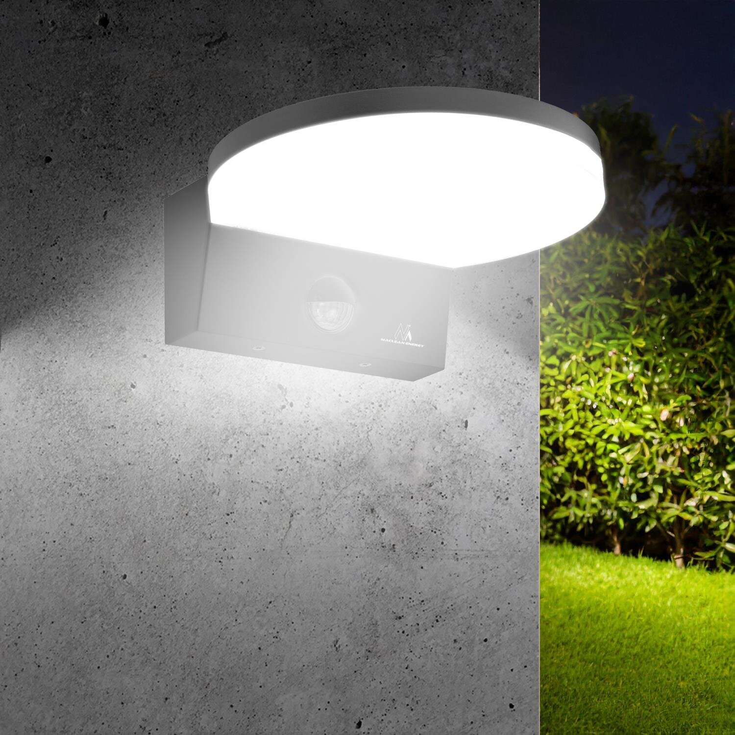 Na obrazie widoczna jest lampa LED zamontowana na betonowej ścianie, emitująca jasne, białe światło. W tle znajduje się zielony trawnik i krzewy.