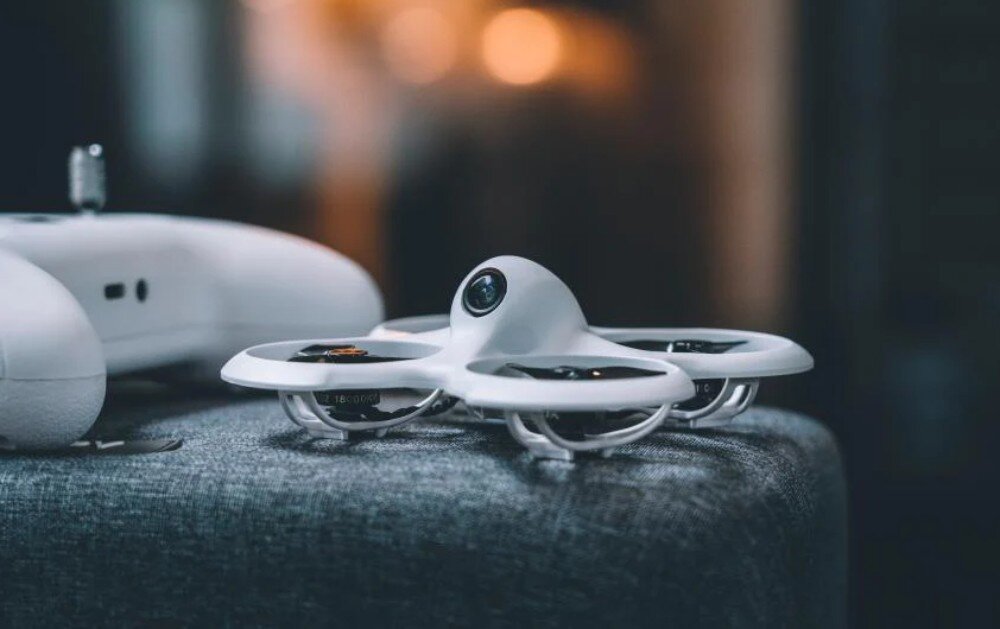 Dron BETAFPV Cetus Pro FPV Kit Urządzenie latające ustawione na szarej materiałowej powierzchni z rozmytym zarysem kontrolera widocznym w tle