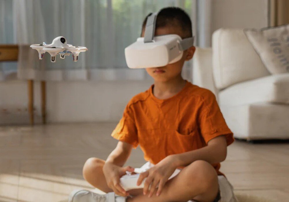 Dron BETAFPV Cetus Lite FPV Kit Dziecko w pomarańczowej koszulce siedzące na podłodze z goglami FPV i sterownikiem, kontrolujące drona