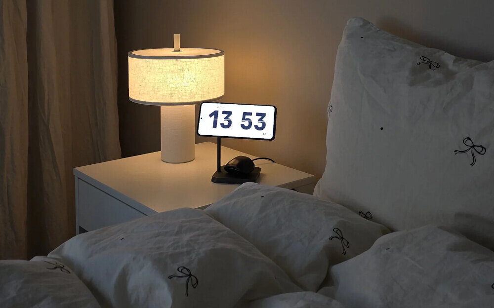 Na stoliku nocnym znajduje się lampa z jasnym abażurem oraz urządzenie z wyświetlaczem pokazującym godzinę 13:53. Obok leży ładowarka indukcyjna z umieszczonym na niej telefonem.
