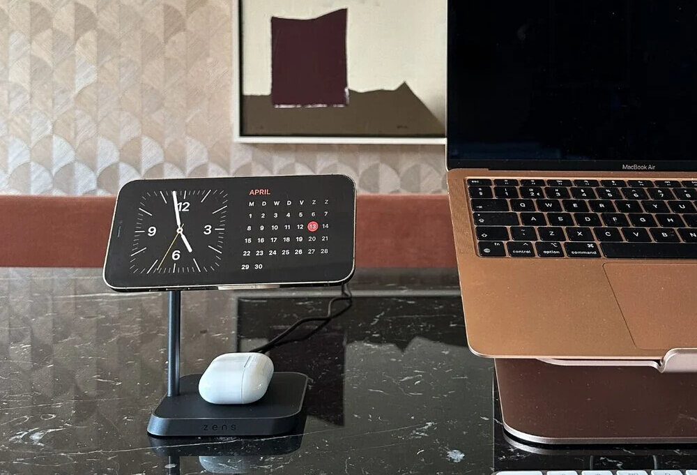 Na marmurowym blacie stoi ładowarka indukcyjna z zegarem i kalendarzem na miesiąc kwiecień, obok znajduje się otwarty laptop. Na podstawce ładowarki leży białe etui na słuchawki, a na froncie podstawki widnieje napis \'zens\'.